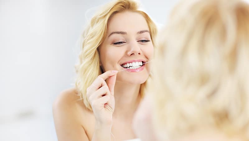 A picture of a young happy woman after a dental cleaning and checkup, flossing her teeth in the bathroom mirror 